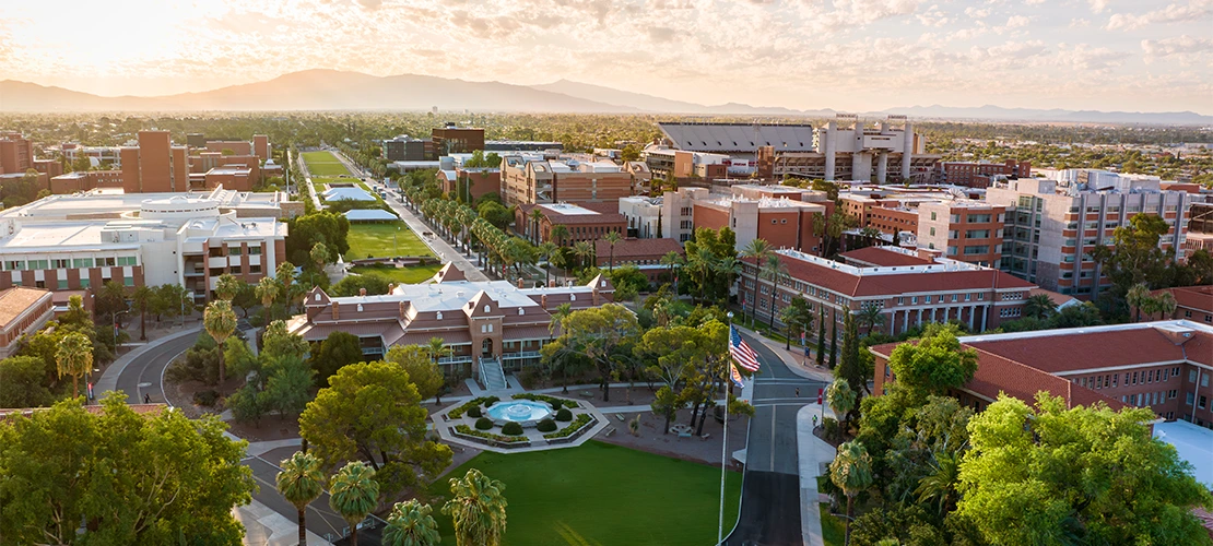 An aerial view of the U of A campus with Old Main in the center, and mountains at sunset in the background.